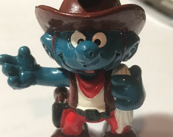 May include: A blue Smurf figurine wearing a brown cowboy hat, red bandana, and brown boots. The Smurf is holding a white cloth in its right hand and a brown gun in its left hand.