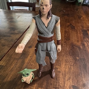 K&ouml;nnte beinhalten: Eine gro&szlig;e Actionfigur einer Figur in einem grau-braunen Outfit, mit braunen Stiefeln und einem braunen G&uuml;rtel. Eine kleine gr&uuml;ne Figur mit gro&szlig;en Ohren und einem beigefarbenen Gewand befindet sich neben der gr&ouml;&szlig;eren Figur. Die Figuren stehen auf einer Holzoberfl&auml;che.