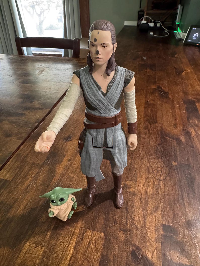 K&ouml;nnte beinhalten: Eine gro&szlig;e Actionfigur einer Figur in einem grau-braunen Outfit, mit braunen Stiefeln und einem braunen G&uuml;rtel. Eine kleine gr&uuml;ne Figur mit gro&szlig;en Ohren und einem beigefarbenen Gewand befindet sich neben der gr&ouml;&szlig;eren Figur. Die Figuren stehen auf einer Holzoberfl&auml;che.