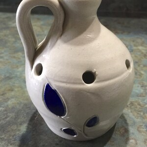 Puede incluir: Una jarra de cer&aacute;mica blanca con un dise&ntilde;o floral azul y agujeros en el lateral. La jarra tiene un asa y un pico.