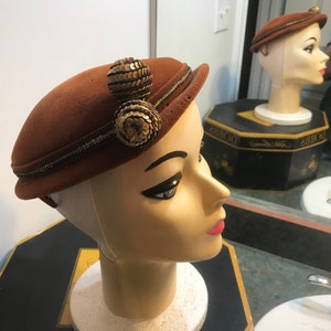 Puede incluir: Un sombrero de fieltro marrón con un adorno de lentejuelas doradas. El sombrero está en una cabeza de maniquí.