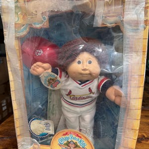 May include: A Cabbage Patch Kids All Stars doll wearing a white baseball uniform with red trim and the word "Cardinals" on the front. The doll is holding a red baseball and is packaged in a box with a yellow and brown background.