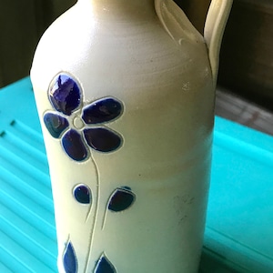 Puede incluir: Una jarra de cer&aacute;mica blanca con un dise&ntilde;o de flor azul. La jarra tiene un asa y un pico.