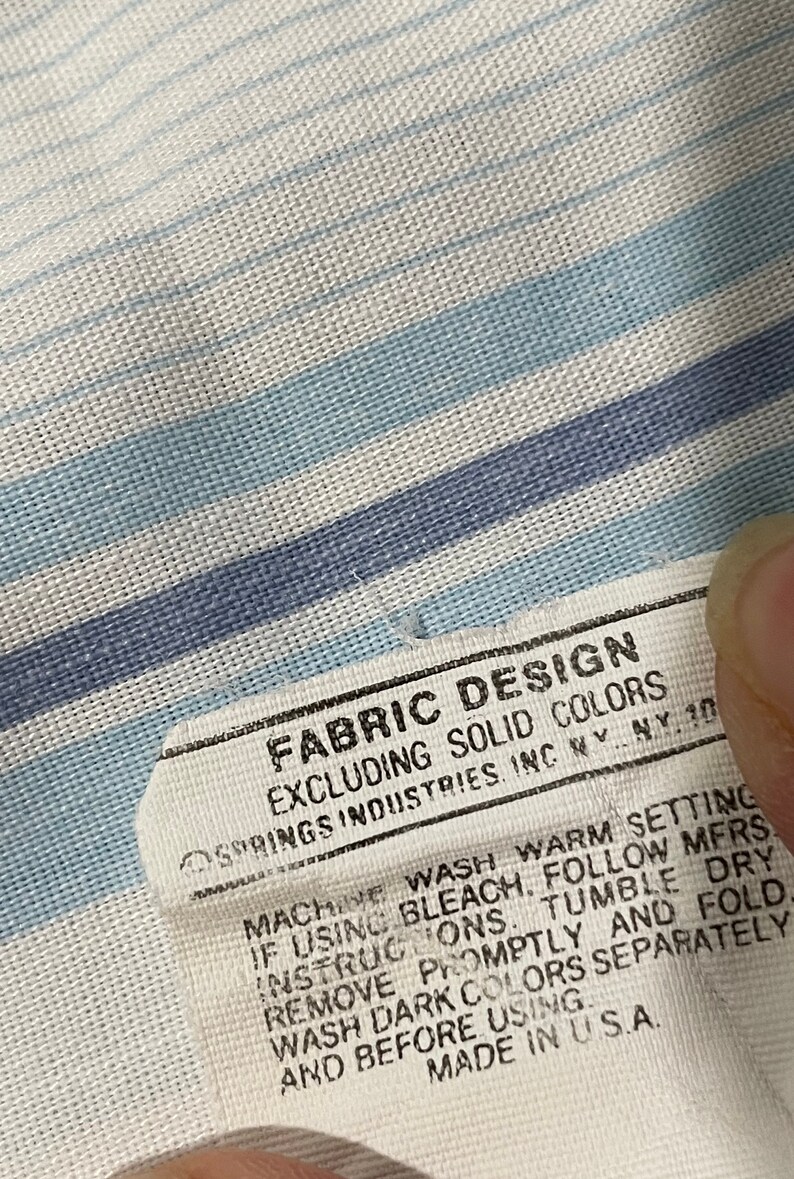 May include: White fabric care label with blue stripes. The label reads "FABRIC DESIGN EXCLUDING SOLID COLORS SPRINGS INDUSTRIES. INC NY NY 10 MACHINE WASH WARM SETTING IF USING BLEACH, FOLLOW MFRS INSTRUCTIONS. TUMBLE DRY REMOVE PROMPTLY AND FOLD WASH DARK COLORS SEPARATELY AND BEFORE USING. MADE IN U.S.A."
