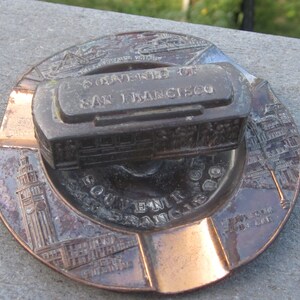 May include: A copper-coloured ashtray with a raised design of the Golden Gate Bridge and the text "University of San Francisco" on a small metal replica of a cable car.