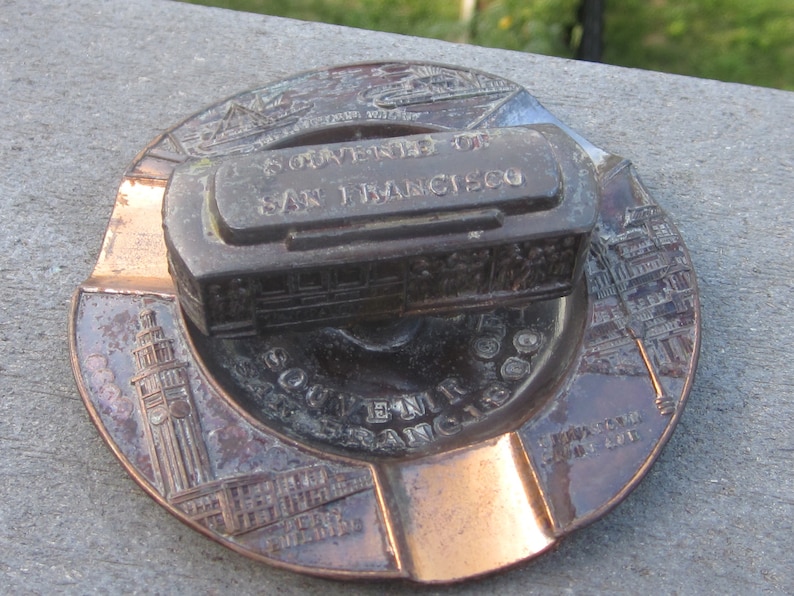 May include: A copper-coloured ashtray with a raised design of the Golden Gate Bridge and the text "University of San Francisco" on a small metal replica of a cable car.