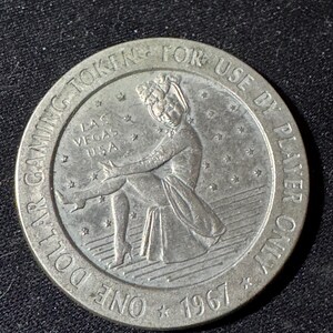 May include: A silver one-dollar gaming token from 1967. The token features an image of a seated woman and the text "LAS VEGAS USA". The outer ring reads "ONE DOLLAR GAMING TOKEN FOR USE BY PLAYER ONLY".