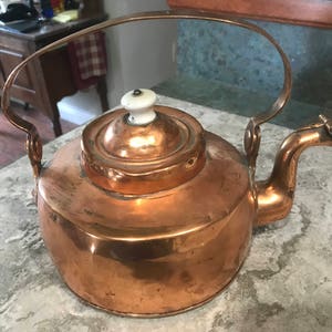 May include: A copper kettle with a rounded body, a curved handle, and a spout. The lid has a white knob. The kettle is sitting on a countertop. The kettle is likely used for boiling water for tea or coffee.