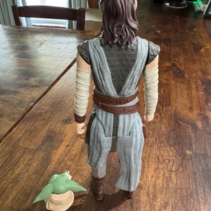 K&ouml;nnte beinhalten: Detaillierte Actionfigur eines Charakters aus einem Science-Fiction-Franchise, die ein grau-braunes Outfit mit Armwickeln und einem braunen G&uuml;rtel tr&auml;gt. Eine kleine gr&uuml;ne Figur mit gro&szlig;en Ohren befindet sich zu F&uuml;&szlig;en der Figur.