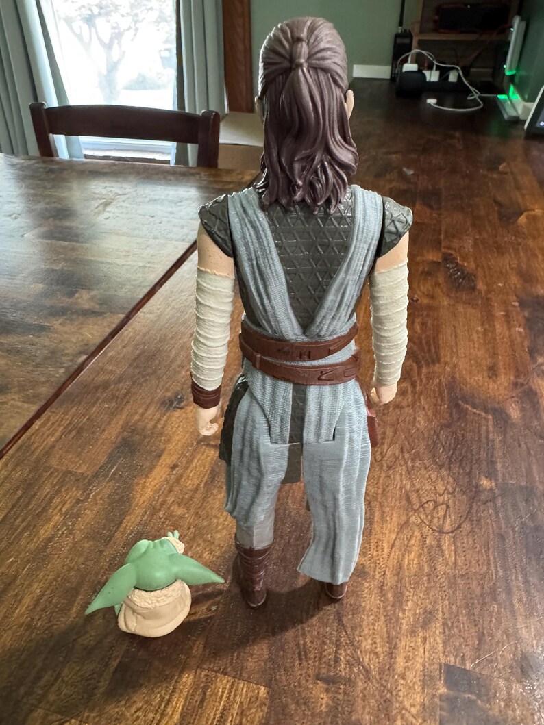K&ouml;nnte beinhalten: Detaillierte Actionfigur eines Charakters aus einem Science-Fiction-Franchise, die ein grau-braunes Outfit mit Armwickeln und einem braunen G&uuml;rtel tr&auml;gt. Eine kleine gr&uuml;ne Figur mit gro&szlig;en Ohren befindet sich zu F&uuml;&szlig;en der Figur.