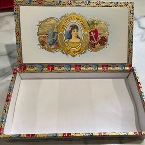 May include: An empty wooden cigar box with a gold and red trim. The lid features a label with a woman's portrait and the text "La Aroma de Cuba Mi Amor".