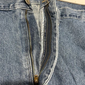 May include: Close-up of a blue denim zip fly on a pair of jeans. The zip is gold metal and the stitching is white.