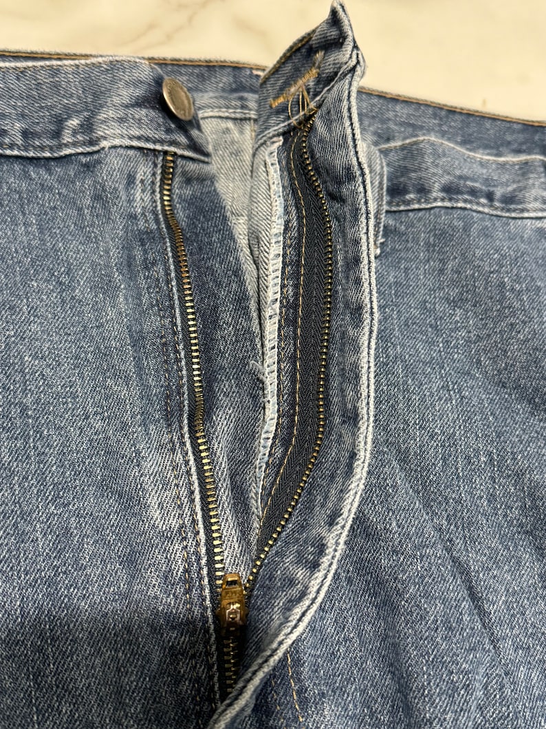 May include: Close-up of a blue denim zip fly on a pair of jeans. The zip is gold metal and the stitching is white.