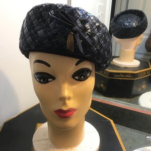 Puede incluir: Un sombrero tejido negro con una cinta negra y un borla de cadena dorada. El sombrero está en una cabeza de maniquí.