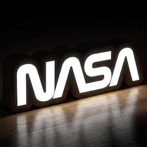 Peut inclure: Un panneau en bois NASA avec des lettres blanches lumineuses. Le panneau est placé sur un fond sombre, les lettres projetant une douce lueur sur la surface en bois en dessous. Les lettres sont en gras et clairement visibles.