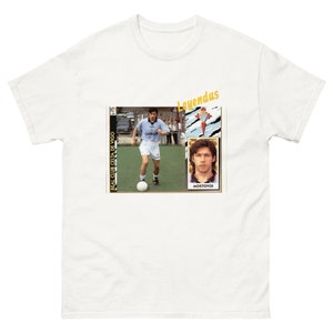 May include: White t-shirt featuring a graphic of a soccer player in a light blue uniform, with the text "Real Club Celta de Vigo." The shirt also includes the word "Leyendas" in yellow and a portrait of a man.