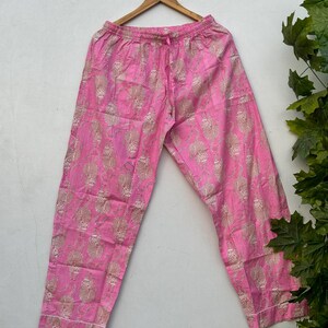 May include: Pink pajama pants with a repeating gold pattern. The pants have a drawstring waist and white trim along the bottom. The pants are hanging on a wooden hanger.