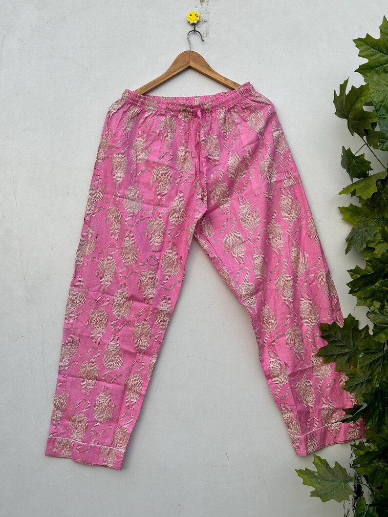 May include: Pink pajama pants with a repeating gold pattern. The pants have a drawstring waist and white trim along the bottom. The pants are hanging on a wooden hanger.