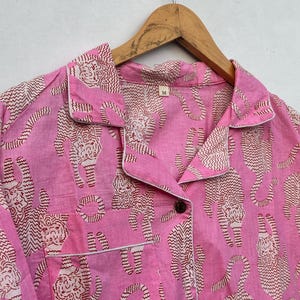 May include: Pink pajama set with a repeating pattern of tigers in a brown and white zig-zag design. The set includes a collared shirt with a breast pocket and white trim, and a button closure. The size tag reads "M".