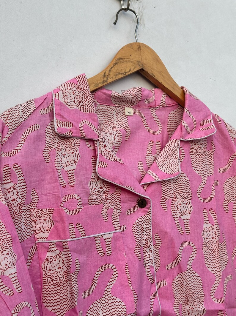May include: Pink pajama set with a repeating pattern of tigers in a brown and white zig-zag design. The set includes a collared shirt with a breast pocket and white trim, and a button closure. The size tag reads "M".