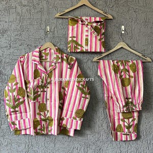 Puede incluir: Un conjunto de pijama a rayas rosas y blancas con un dise&ntilde;o floral. El conjunto incluye una camisa con botones, pantalones y una bolsa con cord&oacute;n a juego. La camisa y los pantalones est&aacute;n colgados en perchas de madera contra una pared gris texturizada.