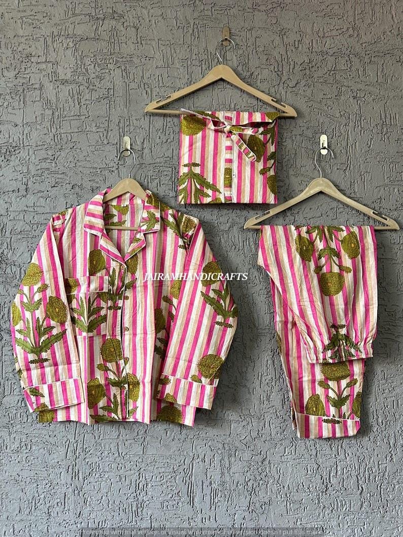 Puede incluir: Un conjunto de pijama a rayas rosas y blancas con un dise&ntilde;o floral. El conjunto incluye una camisa con botones, pantalones y una bolsa con cord&oacute;n a juego. La camisa y los pantalones est&aacute;n colgados en perchas de madera contra una pared gris texturizada.
