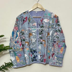 May include: A light blue quilted jacket with a floral and animal print. The jacket features a V-neck and a striped blue and white lining. The text "JAIRAMHANDICRAFTS" is visible on the jacket. The jacket is hanging on a wooden hanger.