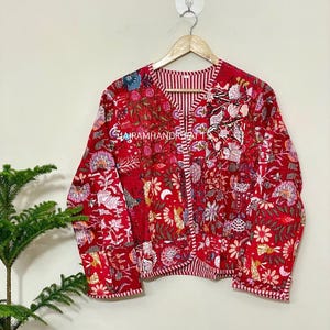 May include: A red quilted jacket with a floral pattern and striped accents. The jacket is hanging on a wooden hanger. The text "CABRAMHANDIGA" is visible on the jacket.