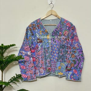 May include: A quilted jacket with a floral pattern in shades of blue, pink, and purple. The jacket has a V-neck and long sleeves. The interior lining is striped in black and white. The jacket is hanging on a wooden hanger.