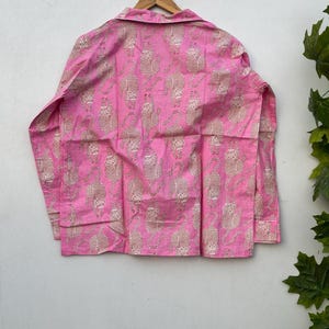 May include: A pink button-down shirt with a repeating pattern of tan tiger illustrations. The long-sleeved shirt is hanging on a wooden hanger against a white wall. The shirt's fabric appears to be lightweight.