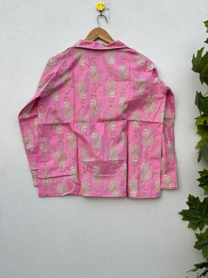 May include: A pink button-down shirt with a repeating pattern of tan tiger illustrations. The long-sleeved shirt is hanging on a wooden hanger against a white wall. The shirt's fabric appears to be lightweight.
