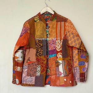 May include: A patchwork jacket in shades of orange, brown, and yellow hangs on a wooden hanger. The jacket features a variety of patterned fabric squares sewn together. The text "Jairam Handicrafts" is visible on the jacket.