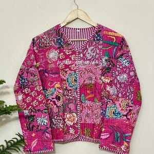 May include: A pink quilted jacket with a floral patchwork design. The jacket has a V-neck and long sleeves. The text "JAIRAMHANDICRAFTS" is visible on the jacket.
