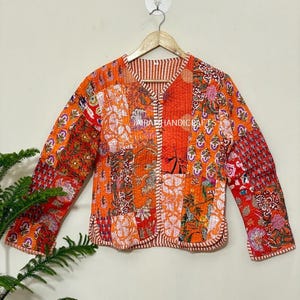 May include: An orange patchwork jacket with floral and geometric patterns. The jacket has long sleeves and a round neckline. The edges are trimmed with a striped fabric. The text "JAIRA HANDICRAFTS" is visible on the jacket.