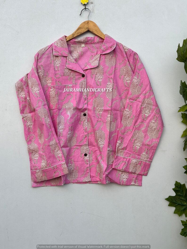 May include: Pink pajama set with a repeating tiger print in a lighter shade. The long-sleeved top has a collared neckline and button-down front. The text "JAIRAMHANDICRAFTS" is visible. The pajamas are hanging on a wooden hanger.