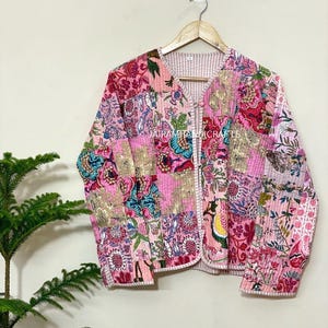 May include: A quilted jacket with a patchwork design featuring floral patterns in shades of pink, gold, and teal. The jacket has a V-neck and is buttoned. The text "JATRAMITABIDICRAFTS" is visible. The jacket is hanging on a wooden hanger.