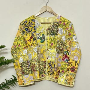 May include: A yellow patchwork jacket with floral patterns and the text "JAIRAMHANDICRAFTS". The jacket has a button-up front and a striped lining. The jacket is hanging on a wooden hanger.
