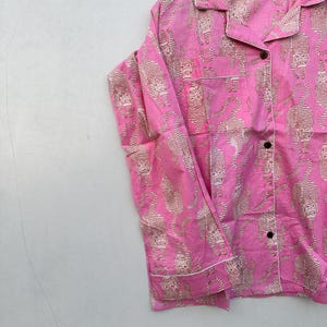 May include: Pink pajama shirt with a repeating tiger print in beige. The shirt has a collared neckline, black buttons, and white trim. The garment is hanging on a wooden hanger.