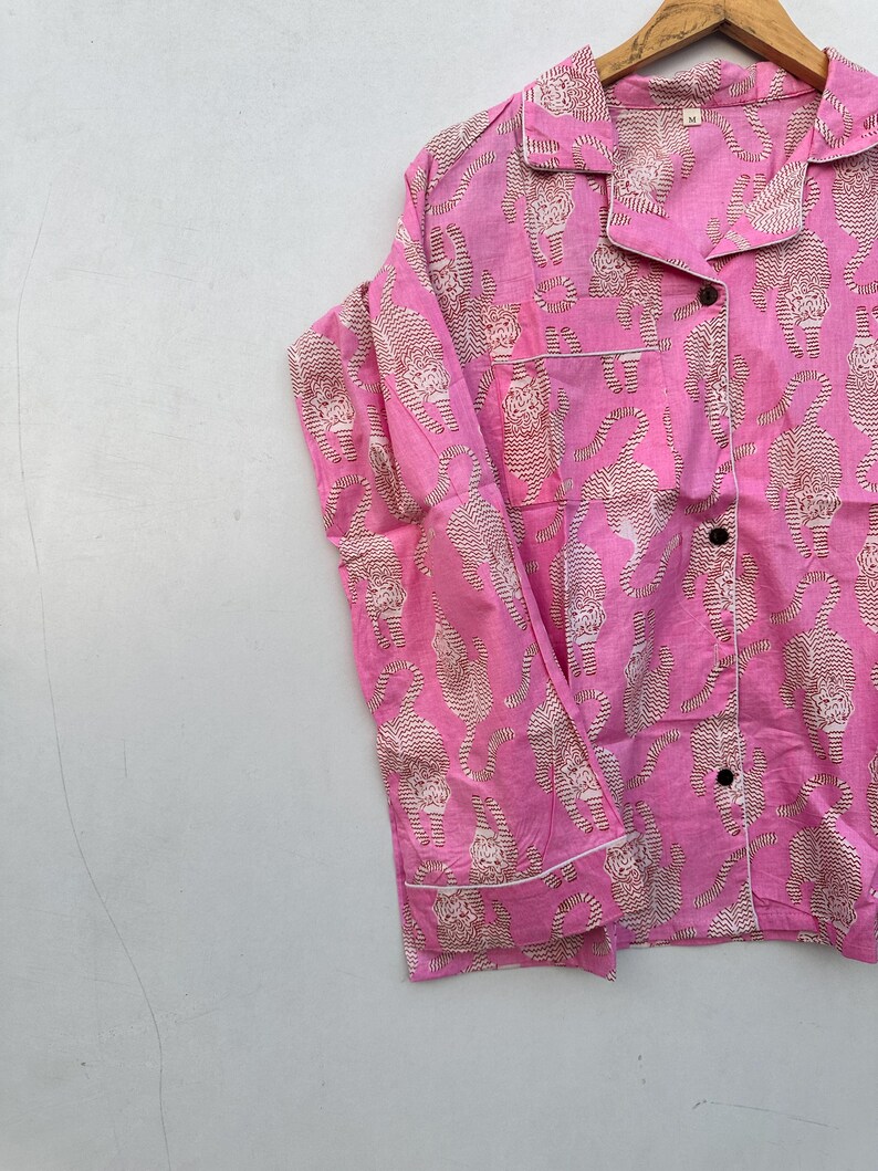 May include: Pink pajama shirt with a repeating tiger print in beige. The shirt has a collared neckline, black buttons, and white trim. The garment is hanging on a wooden hanger.
