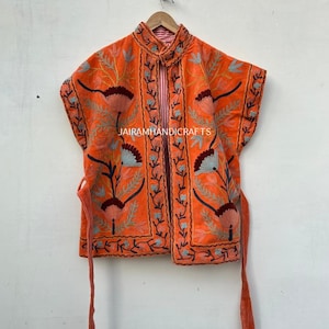 Op de afbeelding: Een oranje geborduurd vest met bloempatronen en een striksluiting. Het vest heeft ingewikkelde borduursels in tinten blauw, bruin en groen. De tekst "JAIRAMHANDICRAFTS" is zichtbaar op het vest.