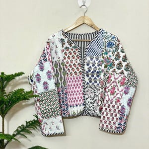 May include: A patchwork jacket with a variety of floral and geometric patterns in shades of pink, green, blue, and white. The jacket has a black and white striped collar and trim, and is hanging on a wooden hanger.