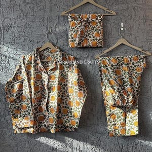 May include: A pyjama set featuring a floral design in yellow, orange, and green on a white base. The set comprises a button-down shirt, trousers, and a matching pouch, displayed on wooden hangers against a textured grey wall.