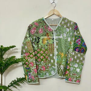 May include: A green quilted jacket with a floral patchwork design. The jacket features a V-neck and is adorned with pink, white, and blue floral patterns. The jacket is hanging on a wooden hanger.