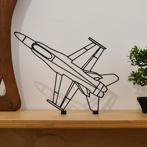 May include: Black metal airplane sculpture. The sculpture depicts a fighter jet with detailed outlines of the wings, fuselage, and tail. The artwork is displayed on a wooden shelf, with a small bonsai tree and a wooden utensil in the background.