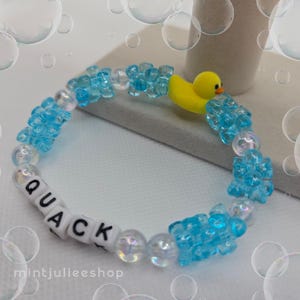 May include: A beaded bracelet featuring translucent light blue flower-shaped beads, iridescent round beads, and white square beads with the black text QUACK. A small yellow rubber duck charm is attached to the top of the circular beaded band.