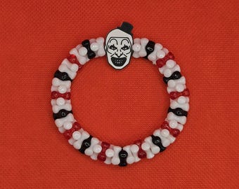 May include: A beaded bracelet with a repeating pattern of red, white, and black beads. A white and black face charm with a top hat is centered at the top of the bracelet. The bracelet is set against a red background.