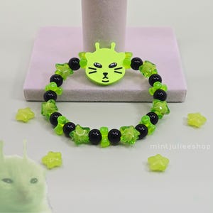 May include: A beaded bracelet featuring alternating black round beads and translucent lime green star-shaped beads. A central lime green charm depicts a stylized cat face with antennae. The text mintjulieeshop is visible in the lower right corner.