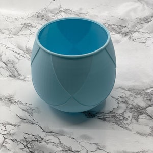 Puede incluir: Una taza de silicona azul claro con un diseño texturizado. La taza está vacía y se encuentra sobre una superficie de mármol blanco.