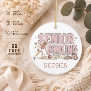 May include: A round white ceramic ornament featuring the text SENIOR BOWLING in pink and blue, with an illustration of a person bowling, a bowling ball, pins, and a bag. The name SOPHIA is printed in pink at the bottom. A gold string is attached to the top.
