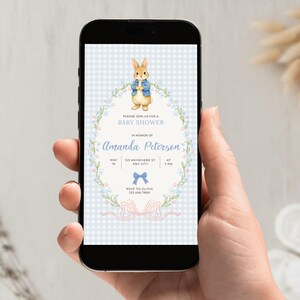 May include: A smartphone displays a baby shower invitation featuring a watercolor illustration of a rabbit in a blue jacket. The invitation includes text, floral accents, and a bow design on a blue gingham background.